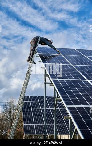 Vertikale Aufnahme von Mann Arbeiter Elektriker Reparatur Photovoltaik-Solarmodul, stehend auf Leiter unter schönen bewölkten Himmel, Installation Solar-Photovoltaik-Panel. Konzept der alternativen Energie Stockfoto