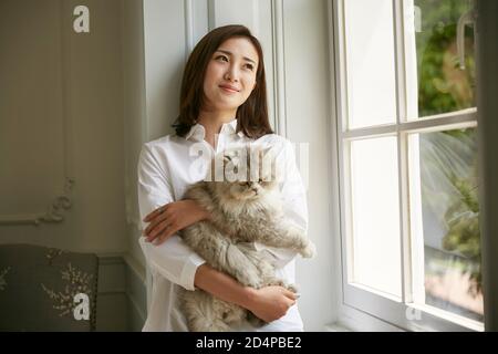 Schöne junge asiatische Frau stehen am Fenster zu Hause halten Eine Katze in den Armen, die gelassen und zufrieden aussieht Stockfoto