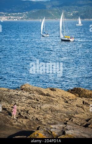PORTONOVO, SPANIEN - 15. AUGUST 2020: Eine junge Frau geht auf den Felsen mit einer Handvoll kleiner Boote, die im Hintergrund entflochten sind. Stockfoto