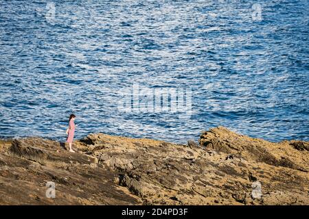 PORTONOVO, SPANIEN - 15. AUGUST 2020: Eine junge Frau überprüft ihr Smartphone und genießt den Sonnenuntergang auf den Felsen in den Rias Baixas, Galicien. Stockfoto