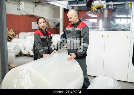 Junge Fabrikarbeiterin und ihr männlicher Kollege stehen bei Riesige verpackte Rolle Stockfoto