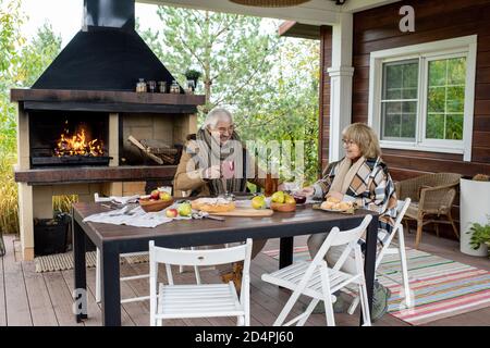 Fröhliches Seniorenpaar in warmer Casualwear mit Tee zum Abendessen Tisch im Freien Stockfoto