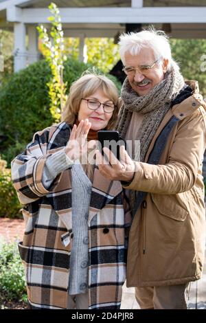 Glückliche Ehegatten im Ruhestand in warmer Casualwear Blick auf ihre Familie Auf dem Smartphone Stockfoto