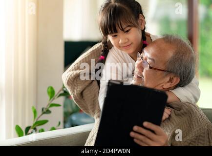 Glücklicher Ruhestand älterer Mann sitzt auf dem Sofa im Wohnzimmer mit Enkelin mit digitalen Tablet zusammen. Familie mit mehreren Generationen und Technologie Stockfoto