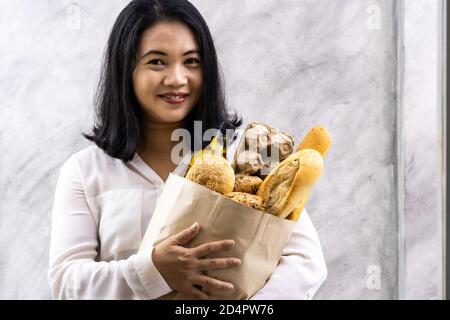 Portrait von asiatischen Lächeln Frau Hausfrau hält Vielfalt Brot in Einweg-Papiertüte auf grau vintage Loft Hintergrund. Bäckerei Essen und Trinken Lebensmittelgeschäft A Stockfoto
