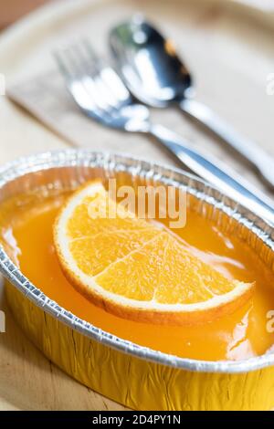 Kuchen mit Stück Orange zum Belag auf Mousse in Aluminiumfolie auf Tisch mit Löffel und Gabel. Beliebte süße Dessert in mit Kaffee servieren Stockfoto