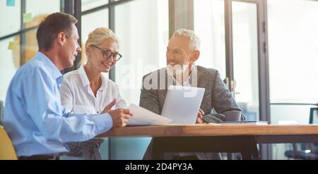Erfolgreiche Geschäftsleute arbeiten zusammen, drei glückliche Mitarbeiter interagieren, diskutieren etwas und lächeln, während am Schreibtisch in der modernen sitzen Stockfoto