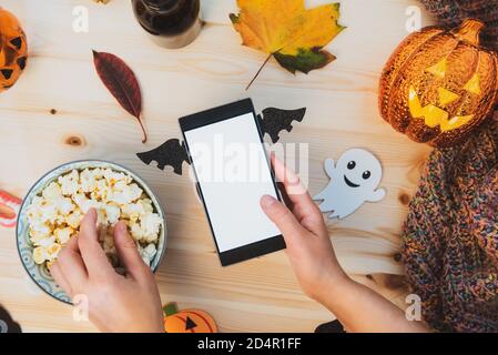 Draufsicht weibliche Hand halten Smartphone mit Fledermaus verziert Flügel mit leerem Bildschirm und Essen Popcorn über Holztisch Mit traditionellem Halloween d Stockfoto