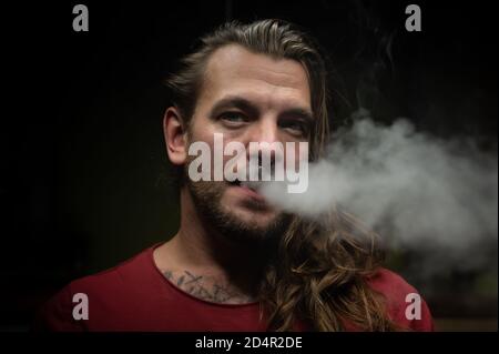 Nahaufnahme eines jungen Mannes, der mit einer elektronischen Zigarette eine Wolke aus Dampf oder Rauch ausatmet. Stockfoto