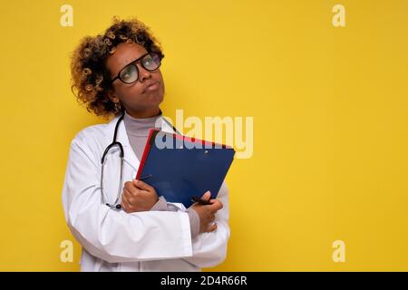 Junger afrikanischer Arzt mit Stethoskop, das Papiere hält und nachdenkt, nach oben zu schauen. Stockfoto
