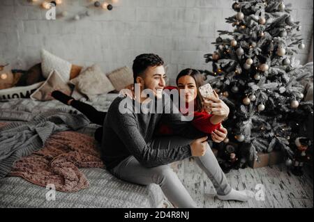 Glückliches junges Paar, das zu Hause Selfie macht, Spaß hat, auf einem gemütlichen Bett zu liegen und neben einem Weihnachtsbaum zu lachen Stockfoto
