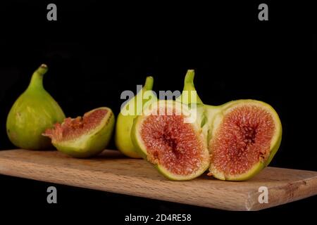 Reife frische Feigen liegen auf einem Holzschneidebrett. Zwei Hälften eines gemeinsamen Feigensykoniums liegen in der vorderen Ebene und zeigen seine eingesäten Früchte. Stockfoto