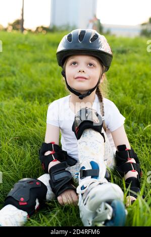 Kleines Mädchen in Schutzausrüstung und Rollen. Sitzen auf dem Gras einen Park. Junge glückliche Skater Stockfoto