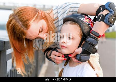Junge Mutter hilft ihrer kleinen Tochter, einen Fahrradhelm anzuziehen. Kinder tragen Sicherheitshemet beim Rollerskaten Stockfoto