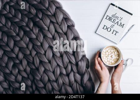 Weibliche Hände halten eine Tasse Kaffee mit Marshmallows auf einem weißen Tisch in der Nähe einer Strickdecke. Stockfoto