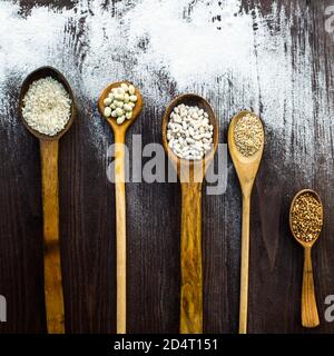 Draufsicht auf Holzlöffel, auf denen verschiedene Getreide auf einem mit Mehl bestreuten Tisch liegen. Stockfoto