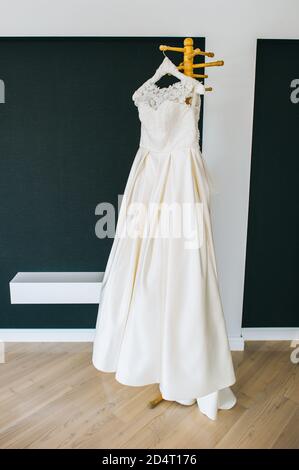 Schönes weißes minimalistisches Hochzeitskleid, das auf einem Holzhänger in der Mitte des Raumes hängt. Stockfoto