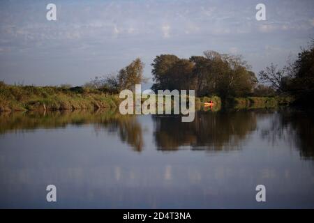 Auf einem Fluss mit dem Herbst gerade beginnt. Es gibt einen Mann in einem orangefarbenen Kanu trägt eine rote Schwimmweste Angeln. Stockfoto