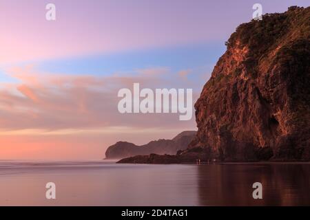 Sonnenuntergang in Piha, Neuseeland. Lion Rock (Vordergrund) ragt über den schwarzen Sandstrand Stockfoto