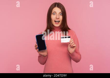 Erstaunt Brünette Frau in rosa Pullover zeigt Handy und Kreditkarte, Blick auf die Kamera mit schockiertem Ausdruck, überrascht mit Online-Banking. Zoll Stockfoto