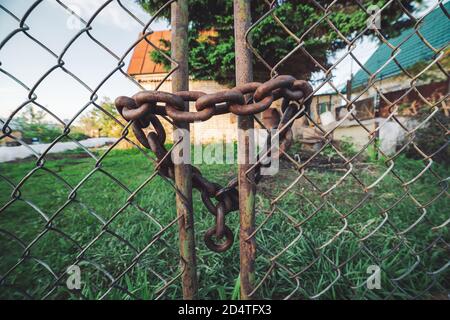 Eiserner rostiger Gitterzaun. Rost auf metallisch verschlossenes Tor. Kette mit Schloss aus nächster Nähe. Grungy Kette in Herzform. Landhaus hinter Stahlgeflecht Stockfoto