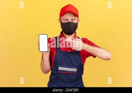 Reparaturman in blauen Overalls rotes T-Shirt und Hut mit Schutzmaske auf Gesicht zeigt Finger auf Smartphone mit leerem Display, Online-Service-Bestellung App Stockfoto