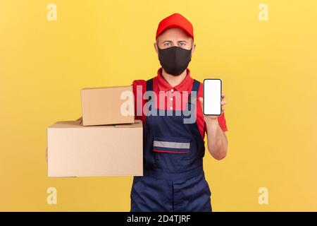 Kurier in Uniform und Schutzmaske auf Gesicht halten zeigt Kartons und Smartphone mit leerem Bildschirm, Anwendung für die Lieferung, Werbung Stockfoto