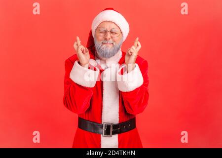 Portrait von verträumten älteren Mann im weihnachtsmann Kostüm Augen schließen und Finger kreuzen einen Wunsch, weihnachtszauber, Winterferien. Studio im Innenbereich Stockfoto