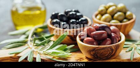 Set aus grünen, roten und schwarzen Oliven und Olivenöl auf grauem Hintergrund. Verschiedene Arten von Oliven in Oliven Holzschalen und Olivenöl auf Holz Schneidebrett und Olivenblätter.Copy space.Horizontal Banner Stockfoto