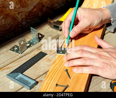 Der Zimmermann versucht mit einem Bleistift an einer Möbelecke. Macht eine Markierung, bevor Sie die Ecke an das Holzbrett Schrauben. Stockfoto