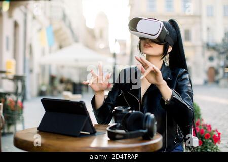 Ziemlich junge intelligente kaukasische asiatische Geschäftsfrau, sitzen im Café im Freien vor dem Tablet, arbeiten und beobachten virtuelle Präsentation in vr-Headset Stockfoto