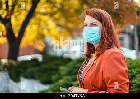 Coronavirus, covid 19 Konzept. Das junge Mädchen in einer medizinischen Maske und Anzug hält ein Smartphone in den Händen und schaut zur Seite auf der Straße Stockfoto