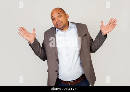 Junge männliche afroamerikanische Geschäftsmann, zuckt, ahnungslos und verwirrt Ausdruck mit erhobenen Armen und Händen. Zweifelskonzept Stockfoto