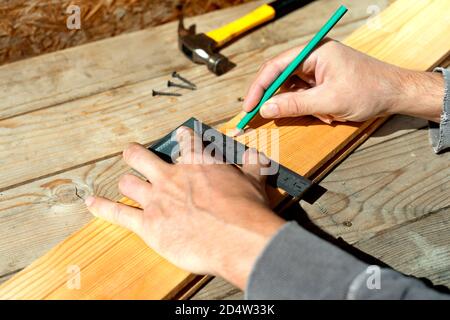 Männlicher Zimmermann markiert mit Bleistift mit einem Quadrat auf dem Brett, Markierungen für Sägeschnitt. Männliche Hände mit Bleistift und einer quadratischen Nahaufnahme auf Holzbrett. Stockfoto