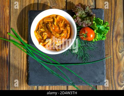 Fisch in Tomatensauce in einer weißen Keramikschale steht auf einem schwarzen Schieferbrett auf einem alten Holztisch. Fisch serviert mit grünen Zwiebeln, Dill, Salat und Tomaten. Snack, Food-Konzept. Blick von oben. Stockfoto