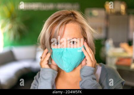 Nahaufnahme einer kaukasischen Frau mit blondem Haar, die im Büro eine medizinische Maske trägt. Sie passt ihre medizinische Maske mit ihren Händen an und sieht erschrocken aus. Geringe Schärfentiefe. Fokus auf Augen. Stockfoto