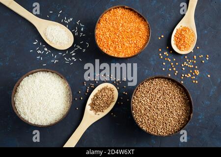 Drei volle Schüsseln mit Cerealien Linsen, Reis und Buchweizen auf dunkelblauem Hintergrund. Die Holzlöffel sind mit Körnern gefüllt. Flache Lay-Komposition. Clo Stockfoto