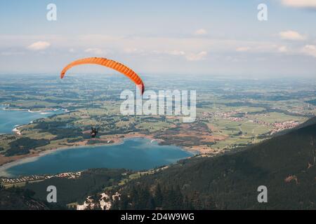 Gleitschirm fliegt von der Tegelbergbahn mit einem orangefarbenen Gleitschirm über Eine schöne Landschaft mit den Seen Bannwaldsee und Forggensee Stockfoto