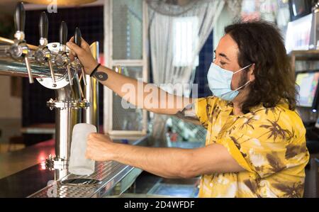 Tätowierte Barkeeper Gießen einen Krug Bier. Er trägt eine medizinische Maske als vorbeugende Maßnahme für die Covid-19-Pandemie. Stockfoto