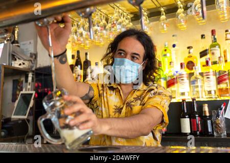 Vorderansicht eines Kellners, der eine Kanne Bier in eine Bar gießt. Er trägt eine medizinische Maske. Stockfoto