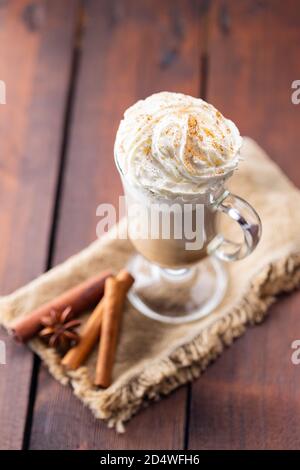 Kürbis Gewürz Latte in einem Glas Becher. Kürbislatte mit Schlagsahne und Gewürzen auf Holzgrund. Heißer Herbstkaffee auf einer Sackleine Stockfoto