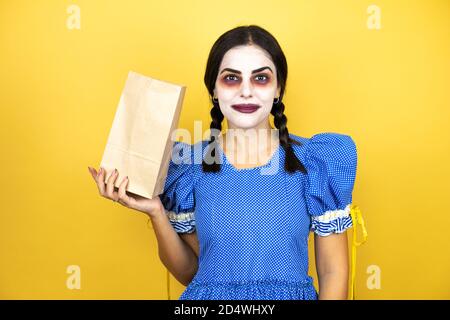 Frau trägt ein gruseliges Puppe halloween Kostüm über gelbem Hintergrund hält eine Papiertasche. Stockfoto