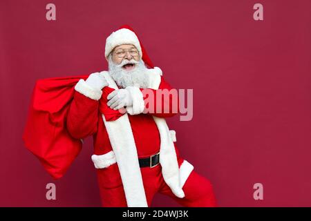 Happy Santa Claus hält Sack mit Weihnachtsgeschenken zu Fuß auf rotem Hintergrund. Stockfoto