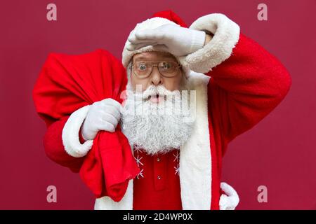 Überrascht schockiert Weihnachtsmann hält Sack mit Weihnachtsgeschenken isoliert auf rot. Stockfoto