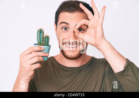 Junger schöner Mann hält kleinen Kaktus Topf lächelnd glücklich tun ok Schild mit Hand auf Auge durch die Finger schauen Stockfoto
