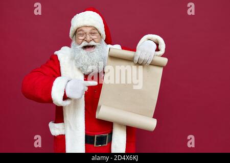 Glücklicher Weihnachtsmann zeigt auf leere Wunschliste isoliert auf rotem Hintergrund. Stockfoto