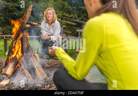 Kaukasische weibliche Freunde in ihren 40er Jahren Hanging vor dem Lagerfeuer Grillen Würstchen und Spaß haben. Camping Kurzurlaub Zeit. Freizeit und Lifestyle Stockfoto