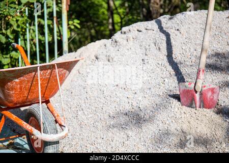 Ein Arbeiter in einem dunkelblauen Overall mit einer Schaufel lädt Sand in eine orangefarbene Baukarre vor dem Hintergrund einer Baustelle. Stockfoto