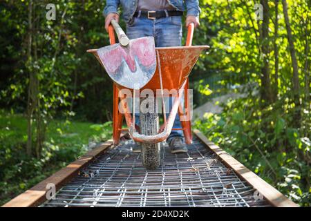 Ein Arbeiter in einem dunkelblauen Overall mit einer Schaufel lädt Sand in eine orangefarbene Baukarre vor dem Hintergrund einer Baustelle. Stockfoto