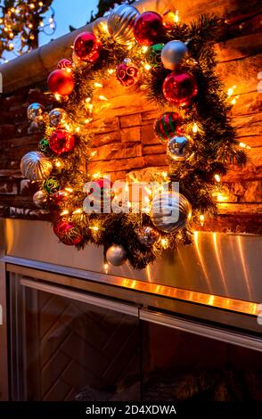 Schöner Weihnachtskranz mit Gold, Silber und Rot mit grünen Ornamenten und Leuchten an einem Kamin im Freien in der Nacht, mit schimmernden Lichtern in der Stockfoto